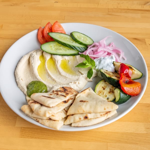 Hummus platter with pita bread, grilled vegetables, tomatoes, cucumbers, pickled onions, and tzatziki.
