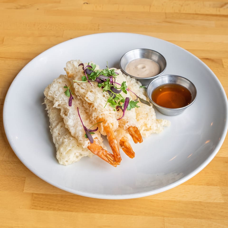 Crispy tempura shrimp garnished with microgreens, served with two dipping sauces on a white plate.