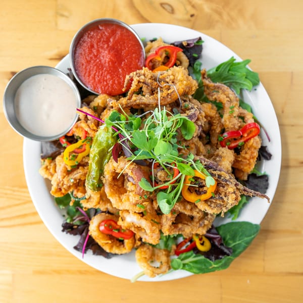 Crispy fried calamari with red and white dipping sauces, garnished with peppers and microgreens.