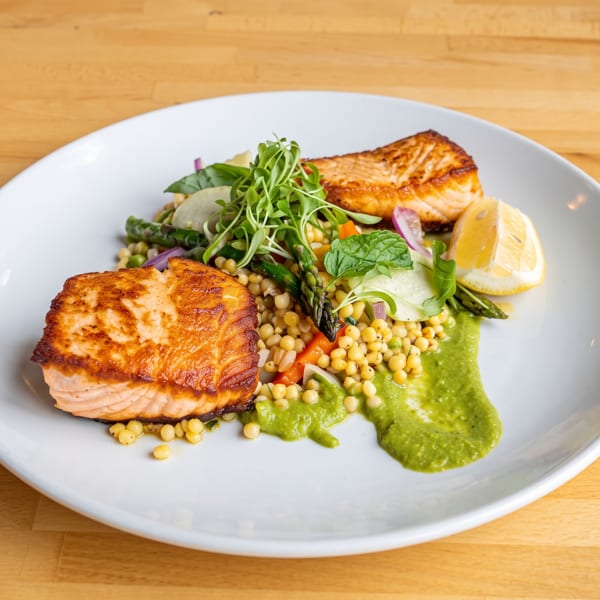 Two salmon fillets with couscous, asparagus, and a green sauce, garnished with lemon and herbs.
