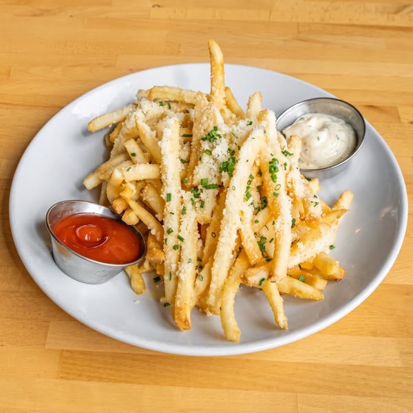 A white plate of golden french fries topped with parmesan cheese and chives, with ketchup and aioli.